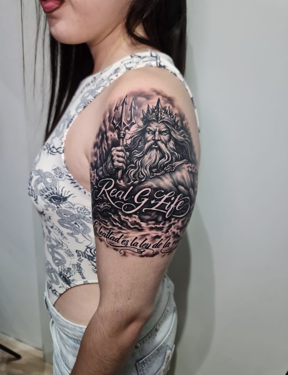 Hombro y parte alta de un brazo tatuados en negro y grises: una figura coronada sostiene un tridente entre nubes y, debajo, dos líneas de letras ornamentales.