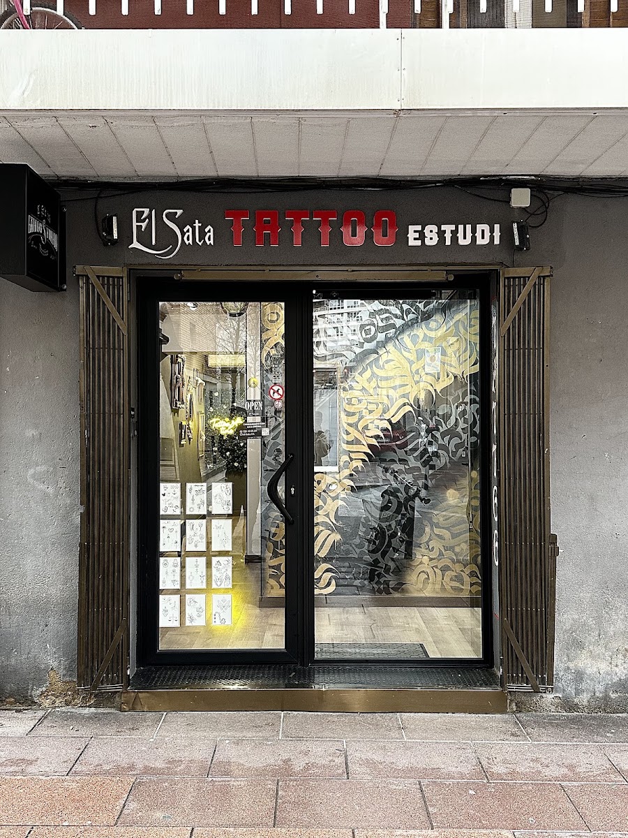 Fachada del estudio en la planta baja: sobre una marquesina gris oscuro se lee El Sata en letra blanca, TATTOO en letras rojas y ESTUDI en blanco. Detrás del cristal, una rejilla de láminas de diseños a la izquierda y un vinilo de letras doradas a la derecha.