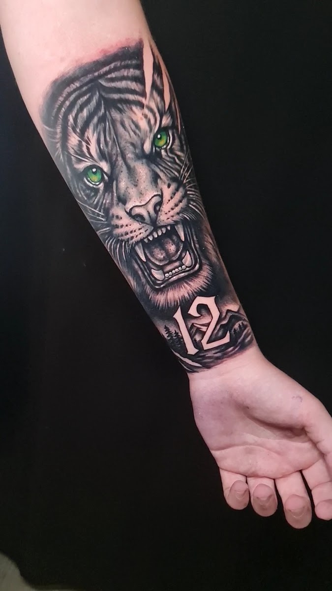 Antebrazo tatuado en negro y grises sobre fondo negro: una cabeza de tigre rugiendo con los ojos verdes y, debajo, una franja de montañas y pinos con el número 12.