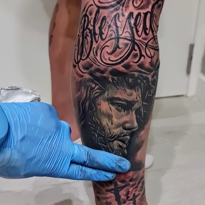 Una mano con guante azul apoyada junto a una pierna recién tatuada en negro y grises: letras ornamentales en la parte alta y, debajo, un rostro barbado con corona de espinas.