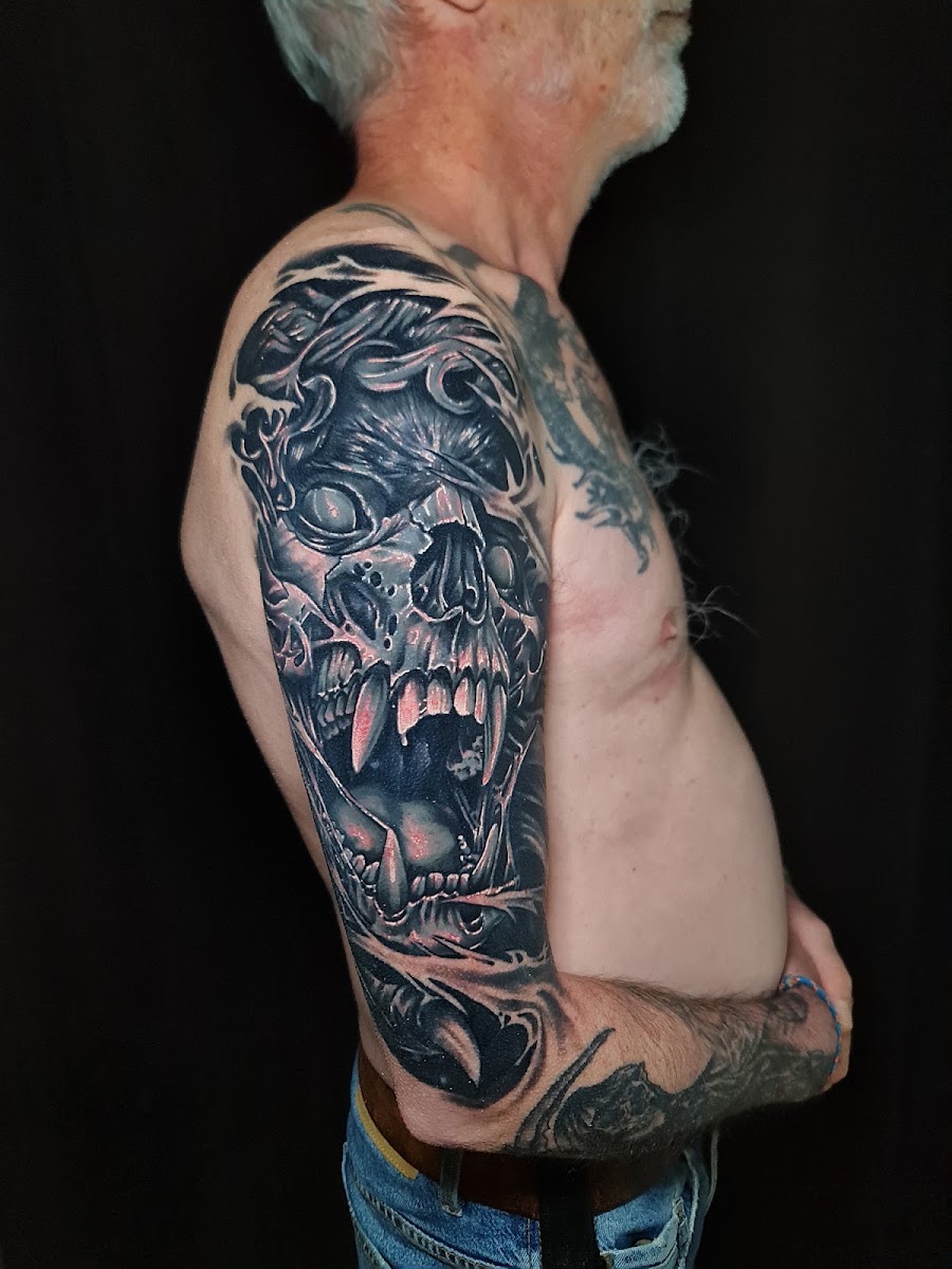 Parte alta de un brazo tatuada en negro y grises sobre fondo negro: un cráneo de fauces abiertas y colmillos largos, rodeado de formas talladas.